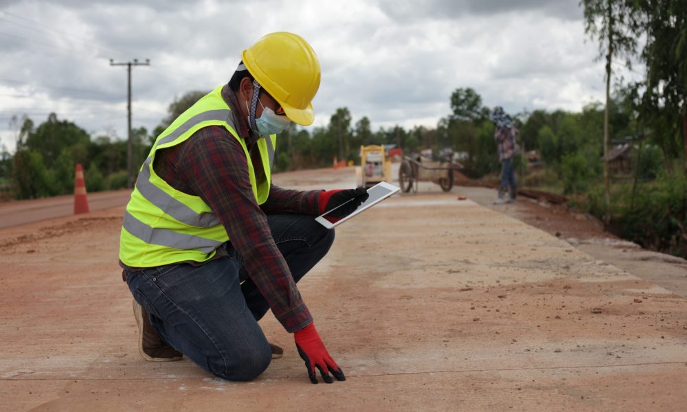 A male contractor wearing a helmet with a tablet bends down to inspect a concrete road at a rural road construction site.